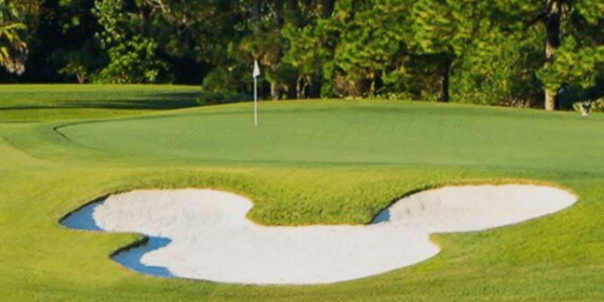 A Mickey Mouse-shaped sand bunker by the green at a Disney World golf course, surrounded by lush trees.