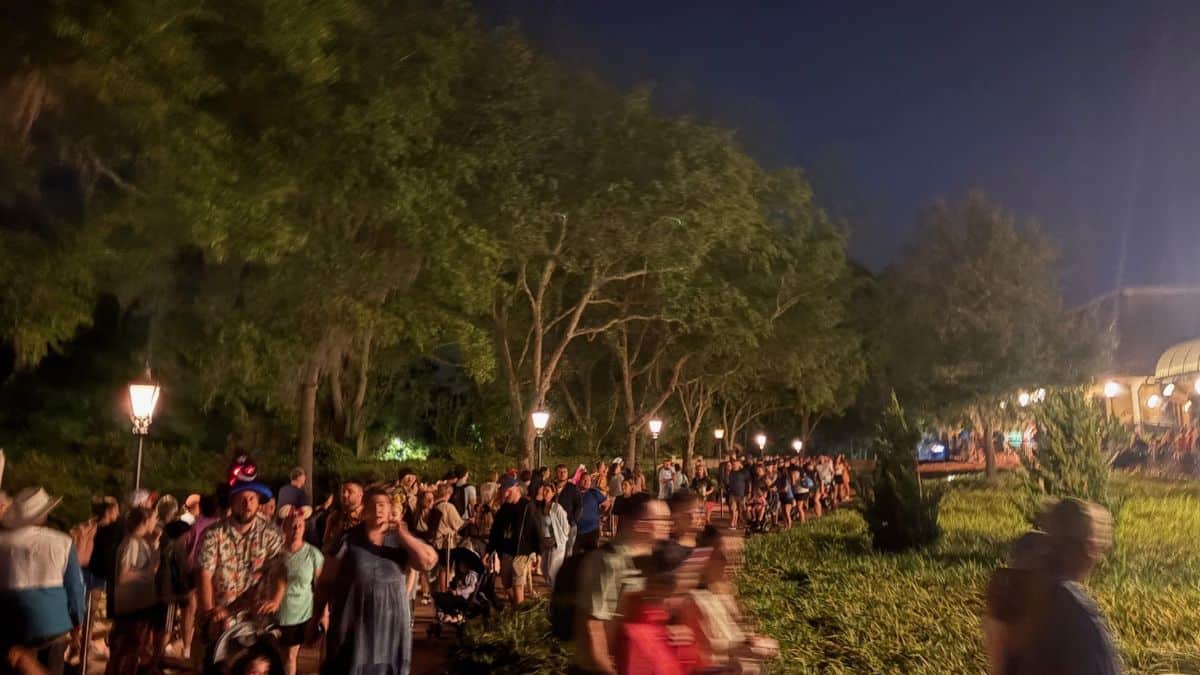 A bustling crowd streams down a lamp-lit, tree-lined path at night—reminiscent of Disney World Skyliner lines at closing time.