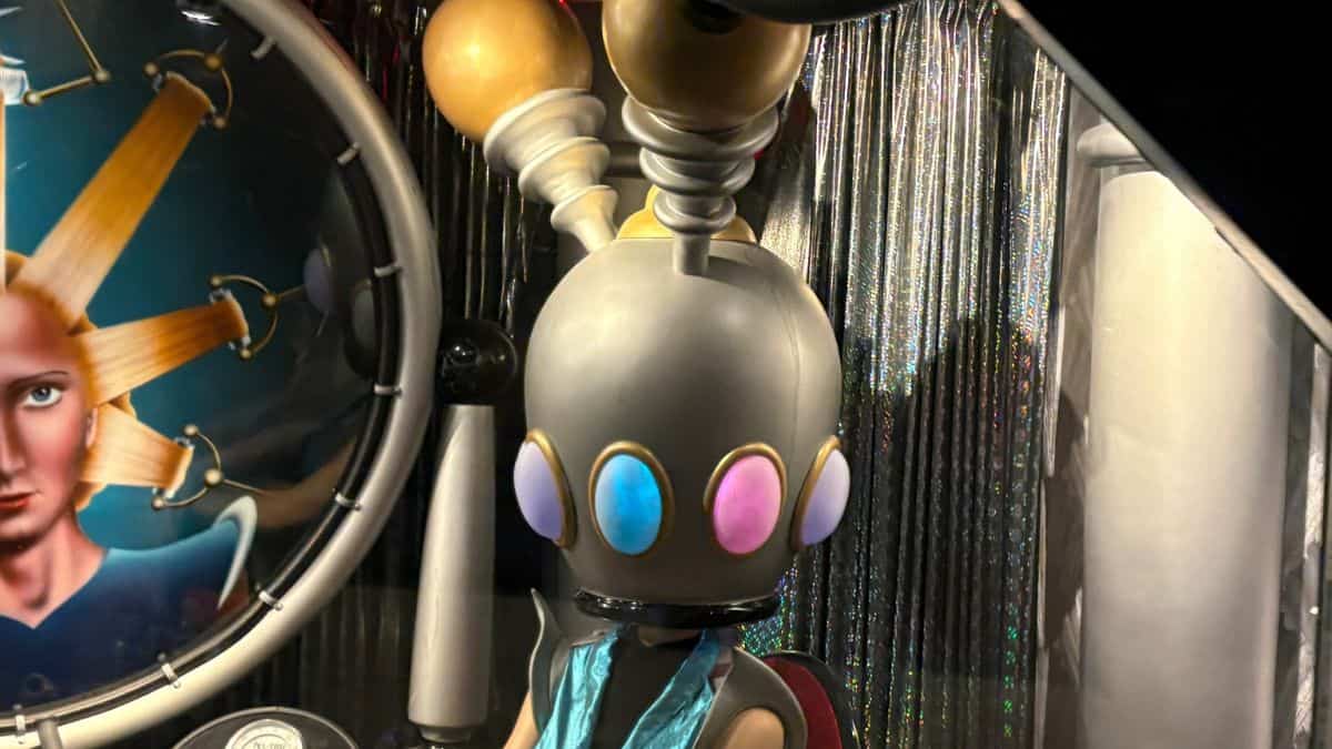 Tomorrowland’s friendly robot mascot, with glowing eyes and antennae, sits by a sleek metallic set in the park.