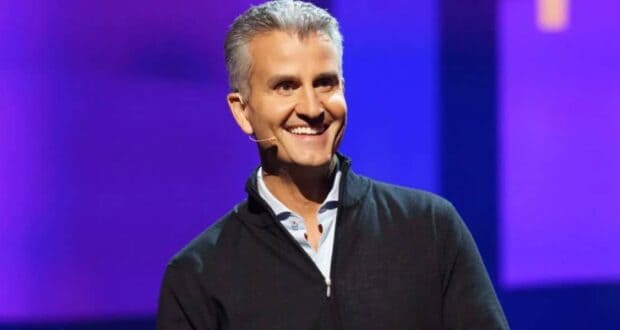 A charismatic host with gray hair and a headset smiles on stage at Epcot, framed by vibrant blue and purple show lights.