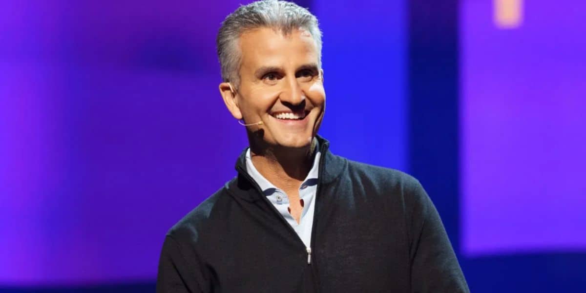 A charismatic host with gray hair and a headset smiles on stage at Epcot, framed by vibrant blue and purple show lights.