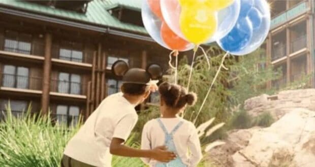 Two children stand together outside, gazing up at a bunch of colorful balloons. The child on the left sports mouse ears, reminiscent of a Disney World guest. Sunlight illuminates the scene, highlighting nearby greenery against the backdrop of balcony-adorned buildings.