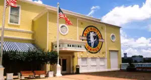 The iconic yellow Atlantic Dance Hall stands out with its bold sign, American flags waving above, inviting benches out front, and a vibrant blue sky dotted with clouds overhead—capturing the classic BoardWalk vibe at Walt Disney World.