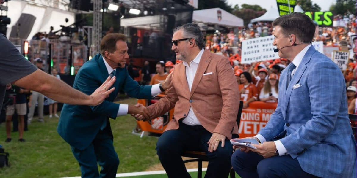 Sports commentators shake hands on the ESPN broadcast stage as enthusiastic fans in the background wave colorful signs, creating a lively, event-like atmosphere reminiscent of theme park parades.