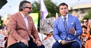 Two sports announcers in sharp suits engage in lively conversation on a broadcast stage, with an enthusiastic audience waving colorful signs and cheering wildly behind them, creating a vibrant atmosphere reminiscent of a high-energy live show you’d find at Universal Studios or ESPN Zone.