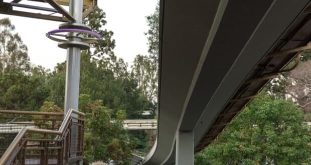 The monorail glides along its elevated track, curving gracefully above lush park trees and a stairway with sleek metal railing, all beneath the signature dramatic atmosphere of an overcast day at the resort.