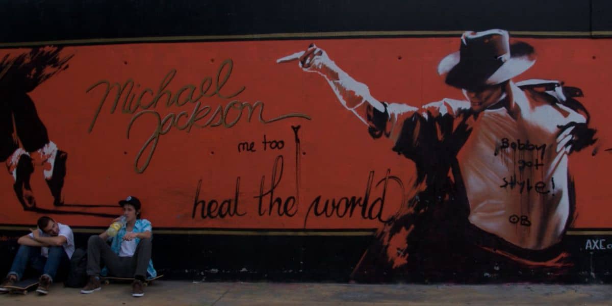Two people sit on skateboards in front of a Universal Studios Michael Jackson mural.