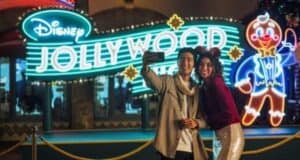 Two guests taking a selfie in front of a Disney Jollywood Nights neon sign at Disney’s Hollywood Studios.