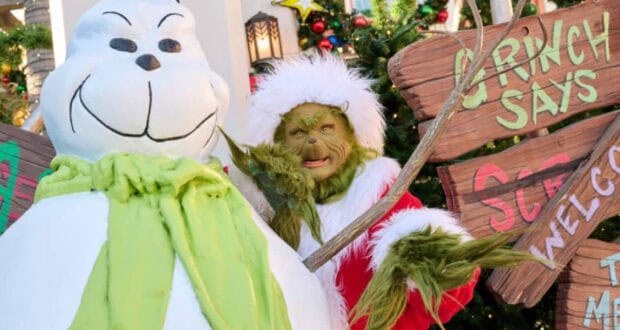 The Grinch in his Santa suit standing by a snowman and Grinch-themed sign at Universal Orlando Resort.