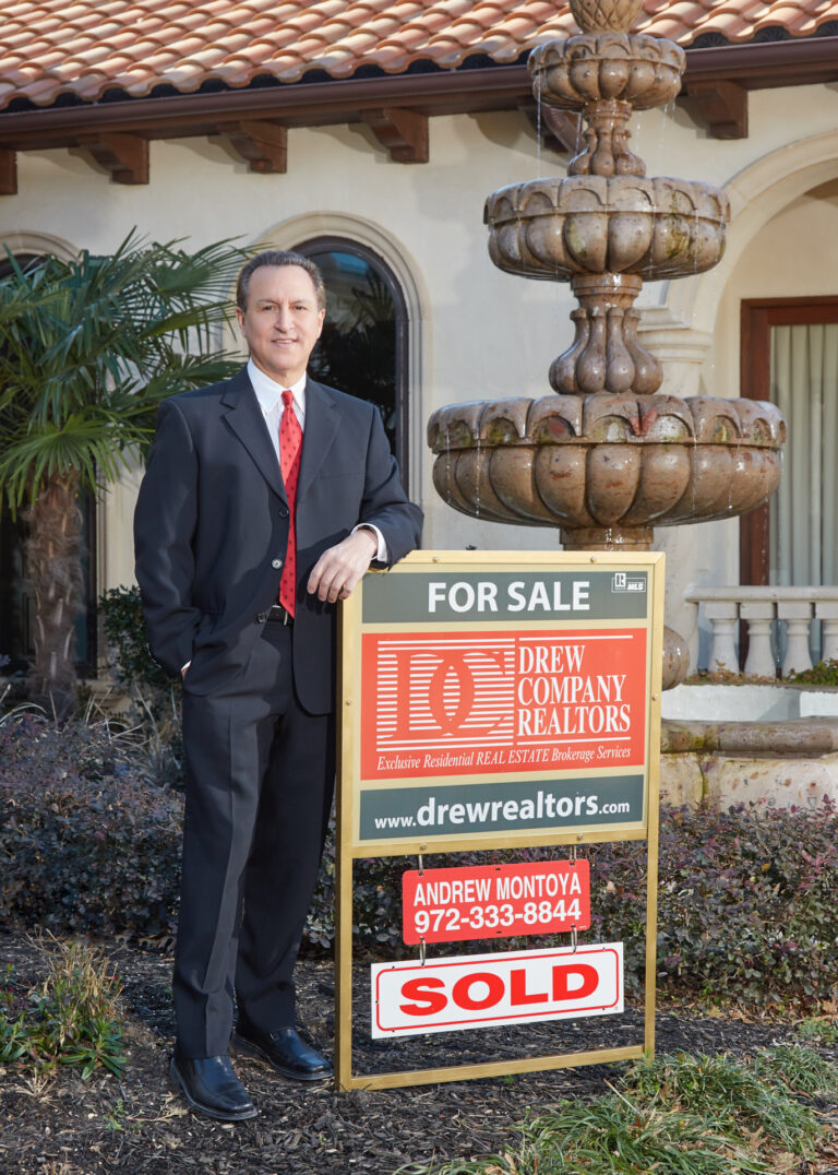 Andrew Montoya standing by a for-sale sign with Drew Company Realtors branding and a fountain in the background.