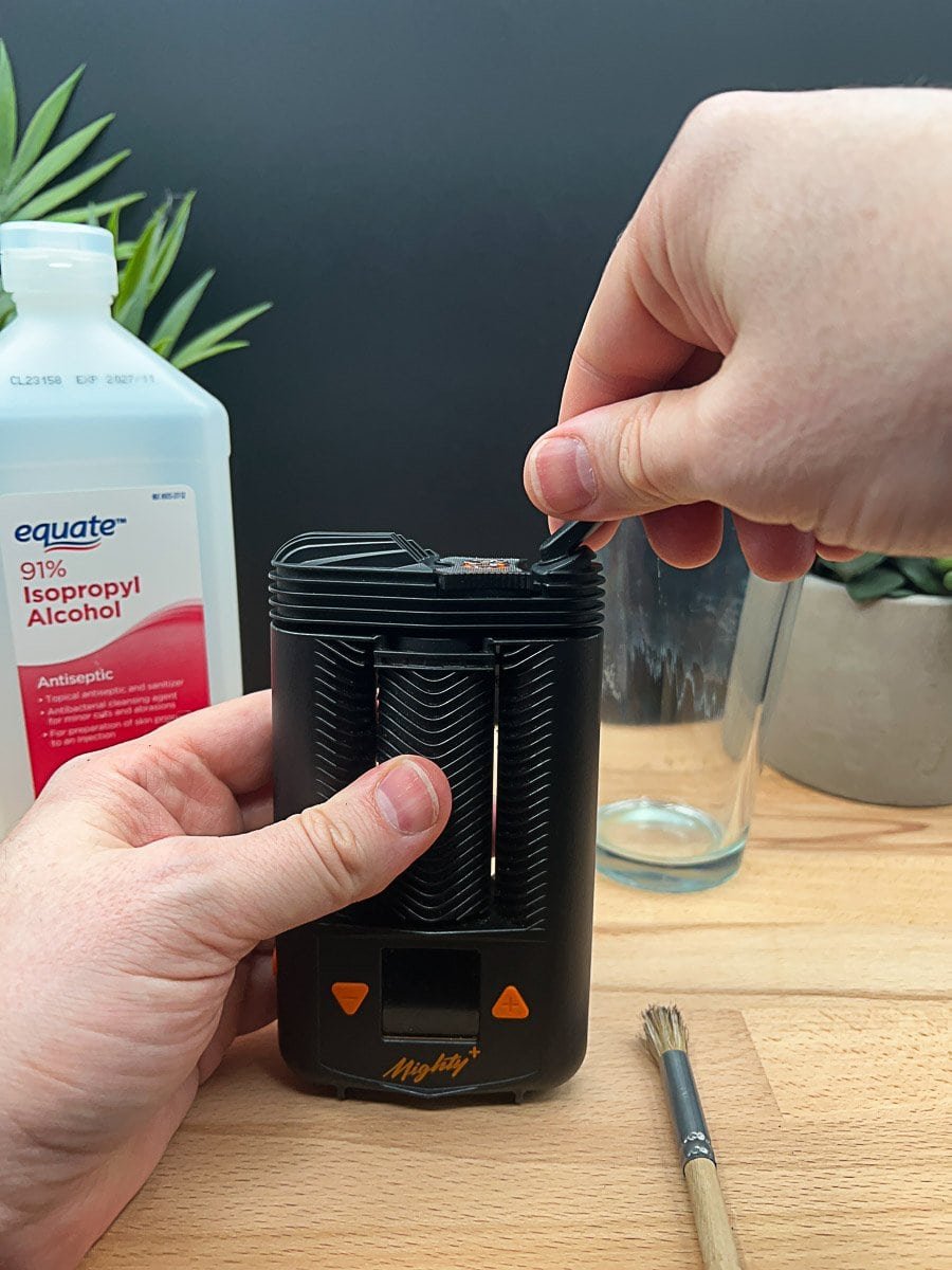 Person cleaning the top of a Mighty+ vaporizer with a brush, with isopropyl alcohol and cleaning supplies visible on a wooden table.