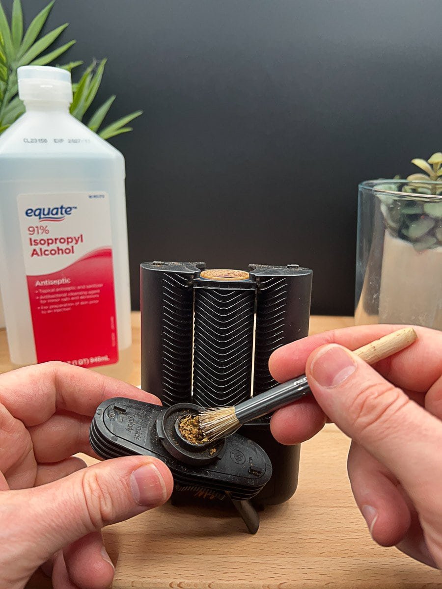 Cleaning a herb grinder with a brush next to a bottle of isopropyl alcohol, with visible residue inside the grinder and plants in the background.