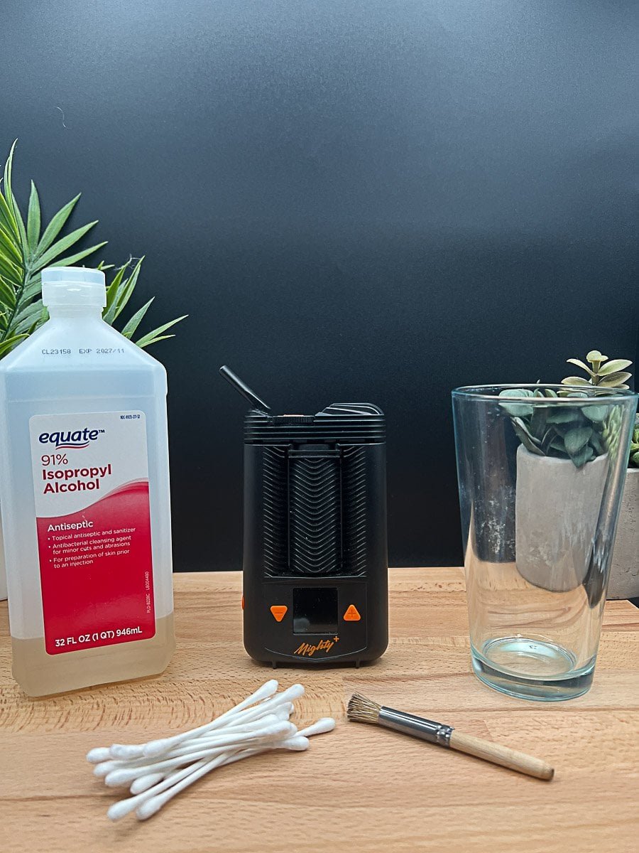 Mighty+ vaporizer cleaning setup featuring isopropyl alcohol, cotton swabs, a small brush, and the device arranged on a wooden table.