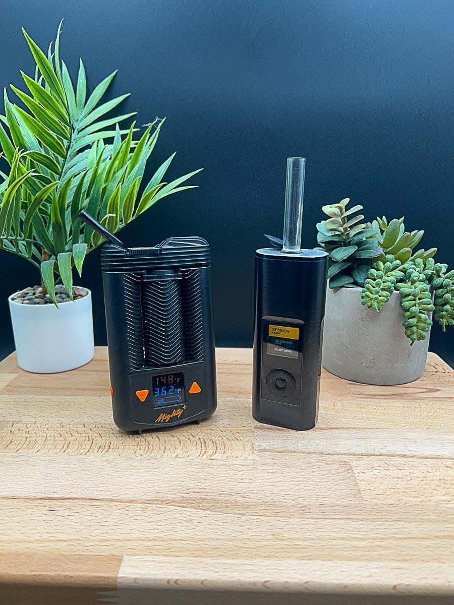 Side‑by‑side comparison of the Mighty+ (Plus) vs Arizer Solo 3 portable vaporizers on a wooden table, showing size, design, and display differences