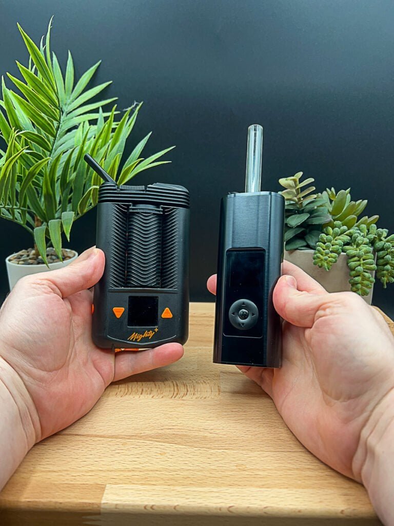 Hands holding the Mighty+ (Plus) and Arizer Solo 3 portable vaporizers side‑by‑side over a wooden table, showing design, size, and interface differences.