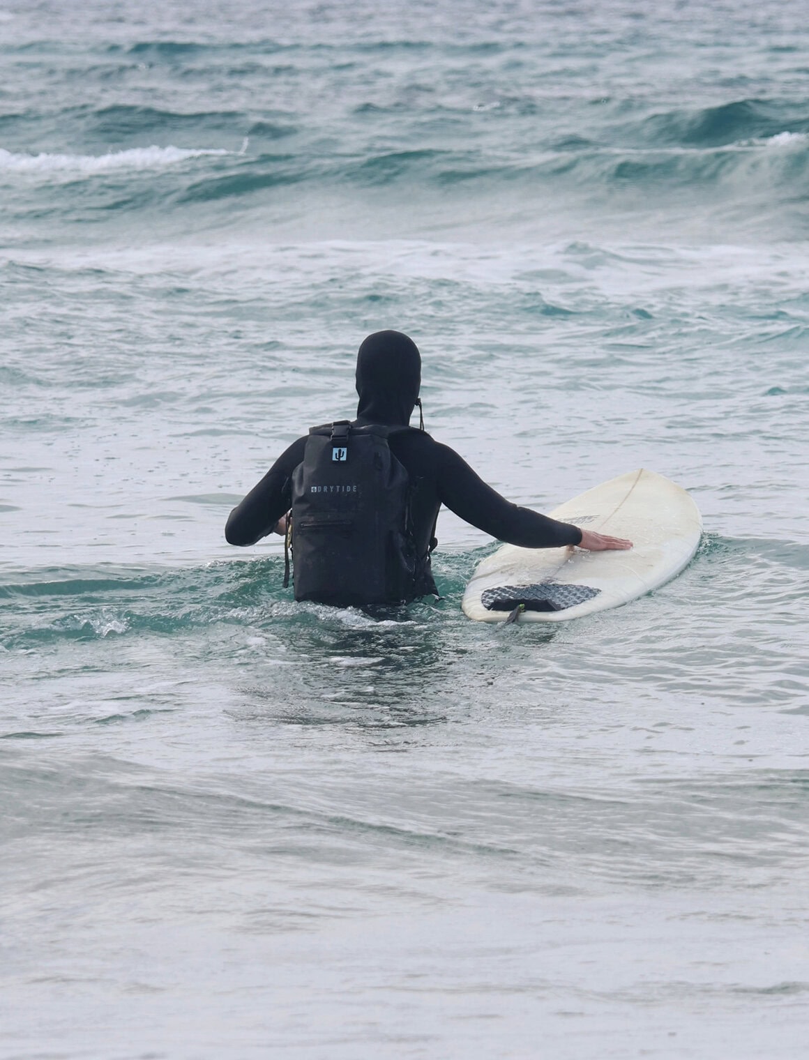 Waterproof backpack user at the beach with surfboard, ocean waves, and overcast sky.