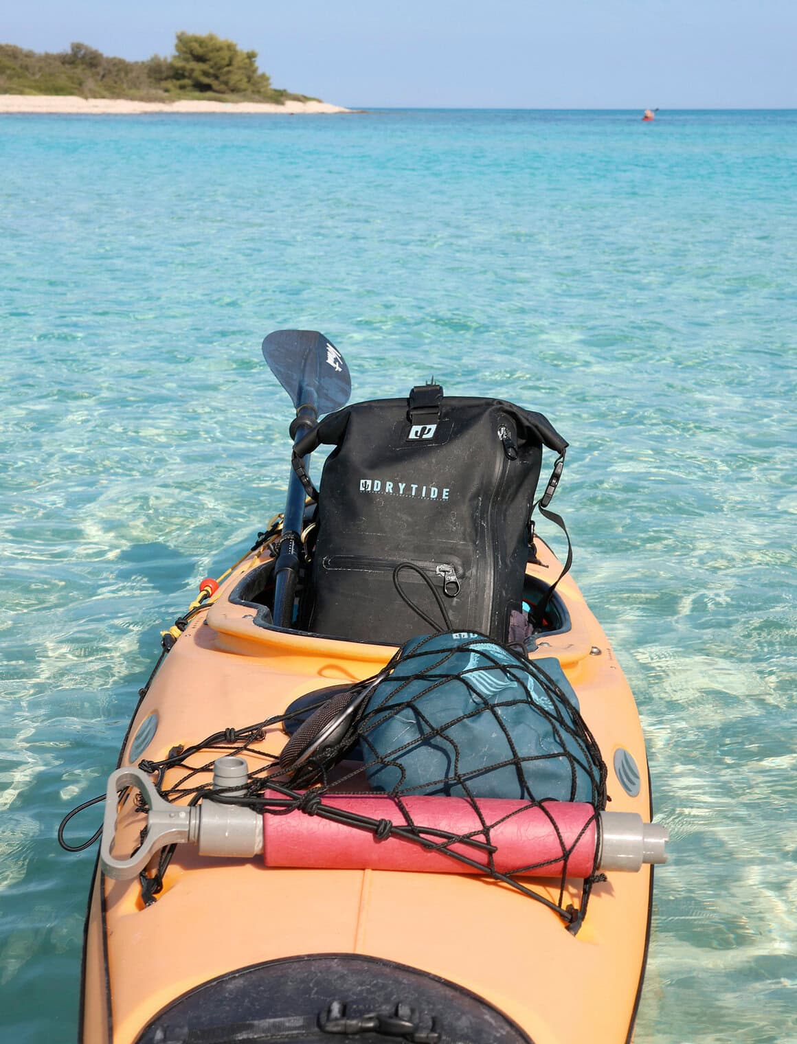 Waterproof backpack on kayak with gear in clear ocean water, sunny sky, and distant shoreline.