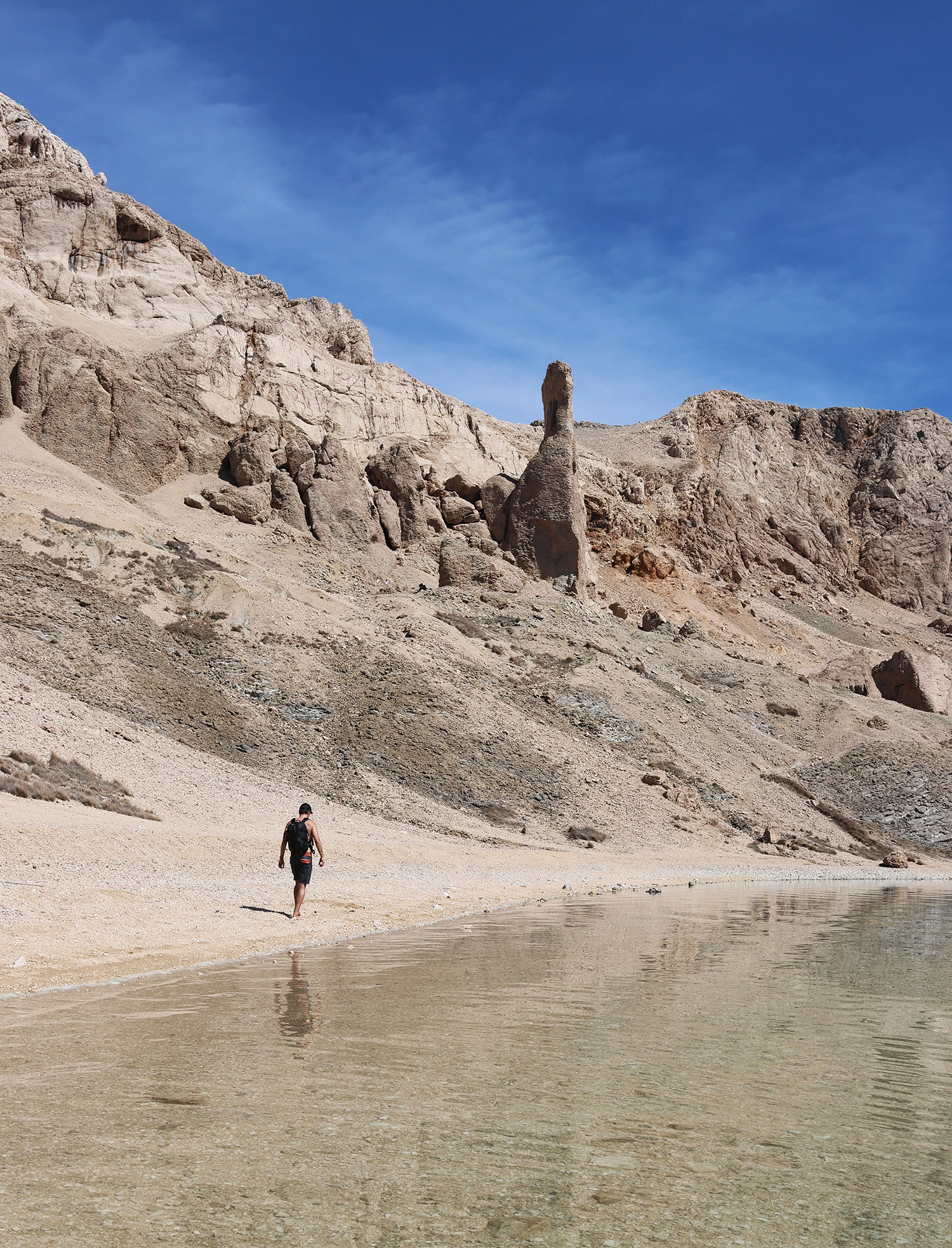 An outdoor hiker walking along a sandy shoreline with rocky cliffs and a clear blue sky in the backg.