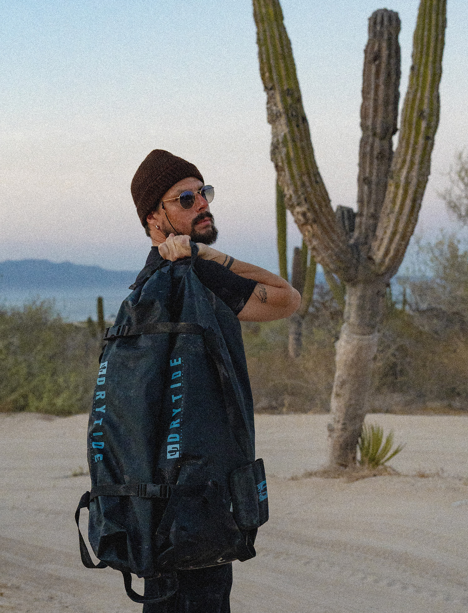 Man with waterproof duffel bag in desert landscape with cactus and mountains.