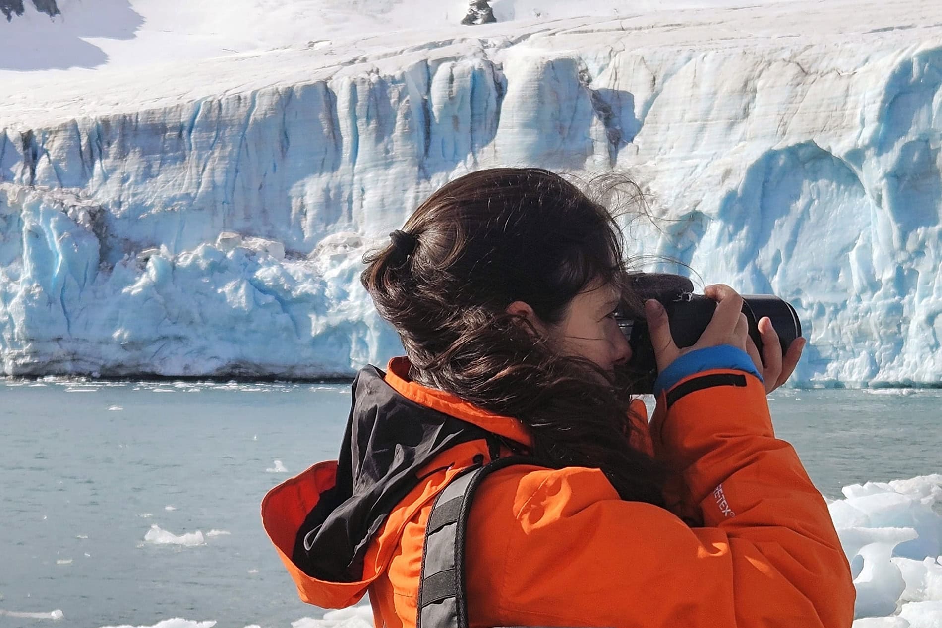 Woman in orange waterproof jacket taking photos in icy landscape.