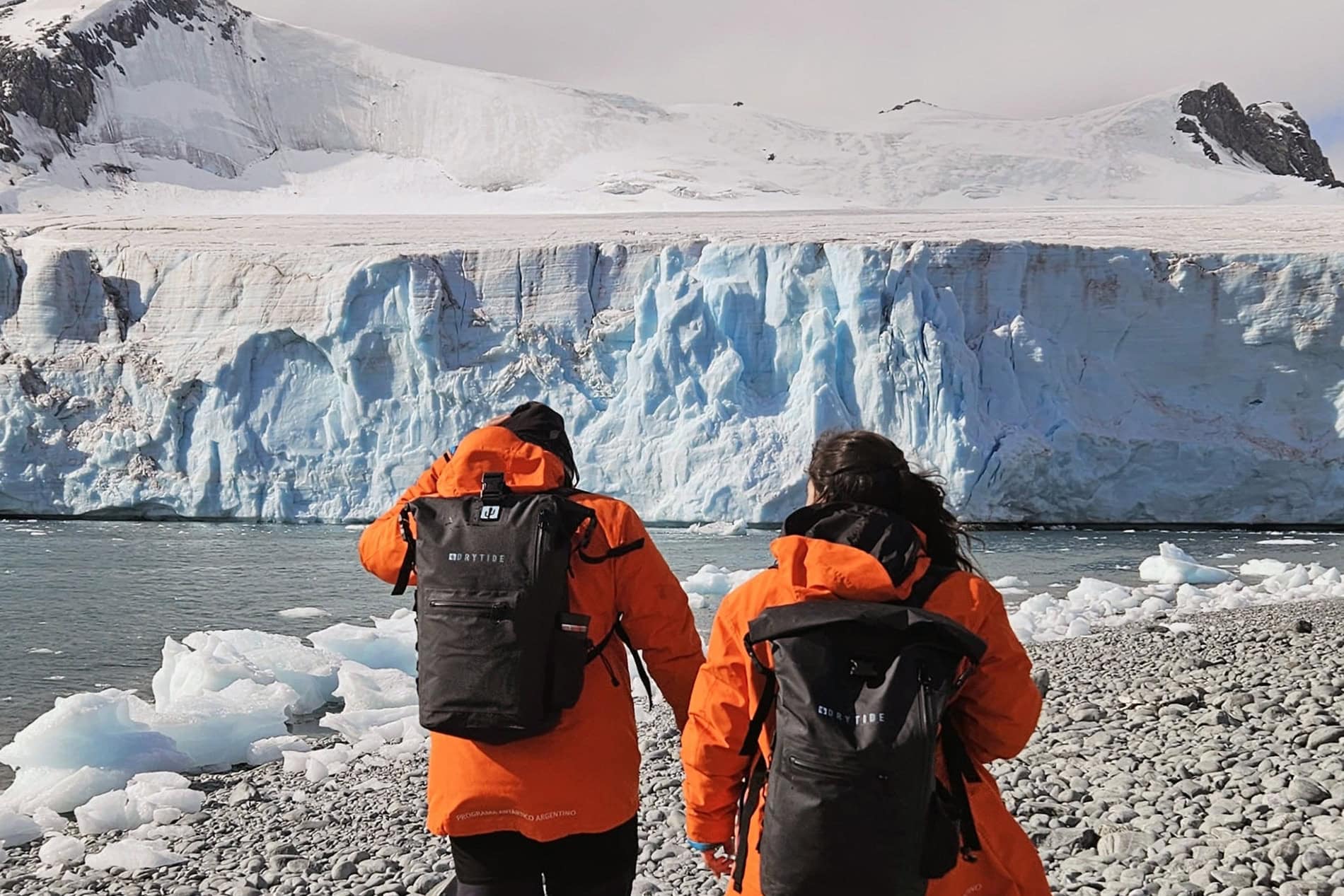 Two hikers in orange jackets with waterproof backpacks near a glacier.