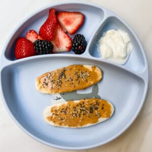 This is a photo of a baby led weaning friendly meal or snack. It contains baby teethers with peanut butter for fat, yogurt for protein and strawberries and blueberries for nutrients and fiber.