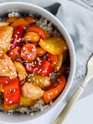 This is a photo of the meal prep friendly chicken stir fry on top of brown rice and topped with sesame seeds in a bowl with a napkin and a fork next to it.