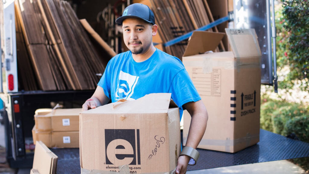 Smiling mover in a blue Element shirt holding an open Element box in a truck