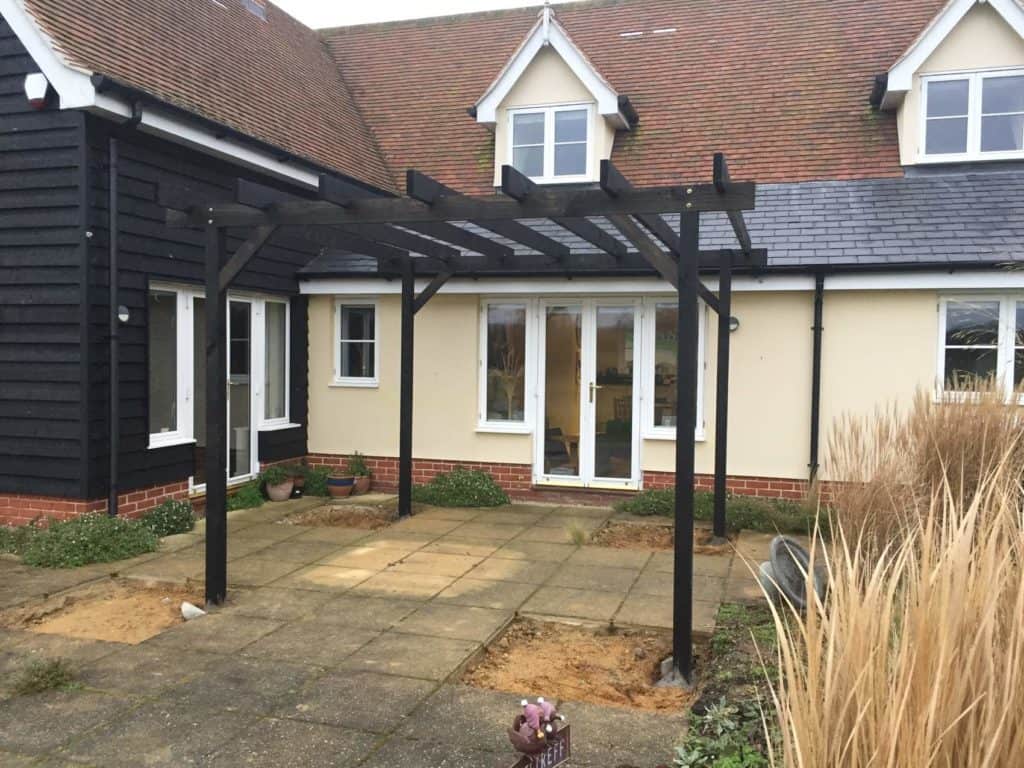Black wooden pergola in a residential garden with paving and house background.
