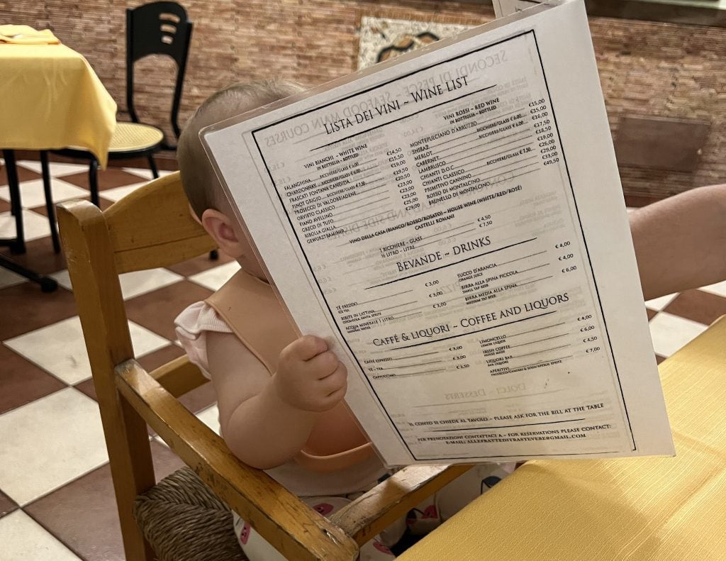 Baby sitting in a high chair holding a menu at a restaurant in Rome, enjoying a family-friendly dining experience with a view of checkered floor and brick wall.