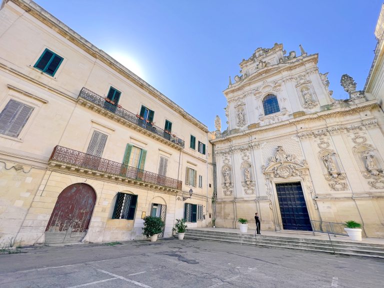 Elegant Baroque church façade in Italy with intricate white stucco details, surrounded by historic buildings, under a clear blue sky, perfect for exploring Italian architecture and travel inspiration.