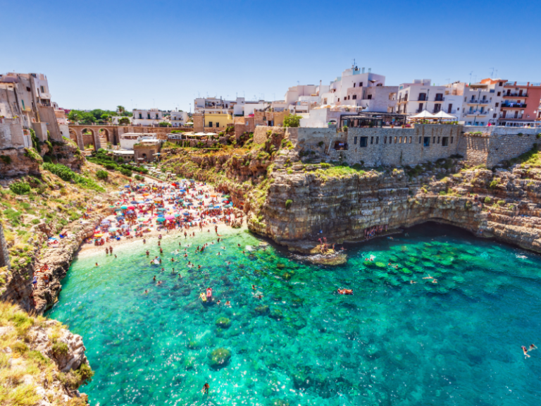 Sunlit Mediterranean coastal cove with turquoise waters, sandy beach, colourful umbrellas, historic cliffs, and charming white-washed buildings, ideal for travel and exploration.