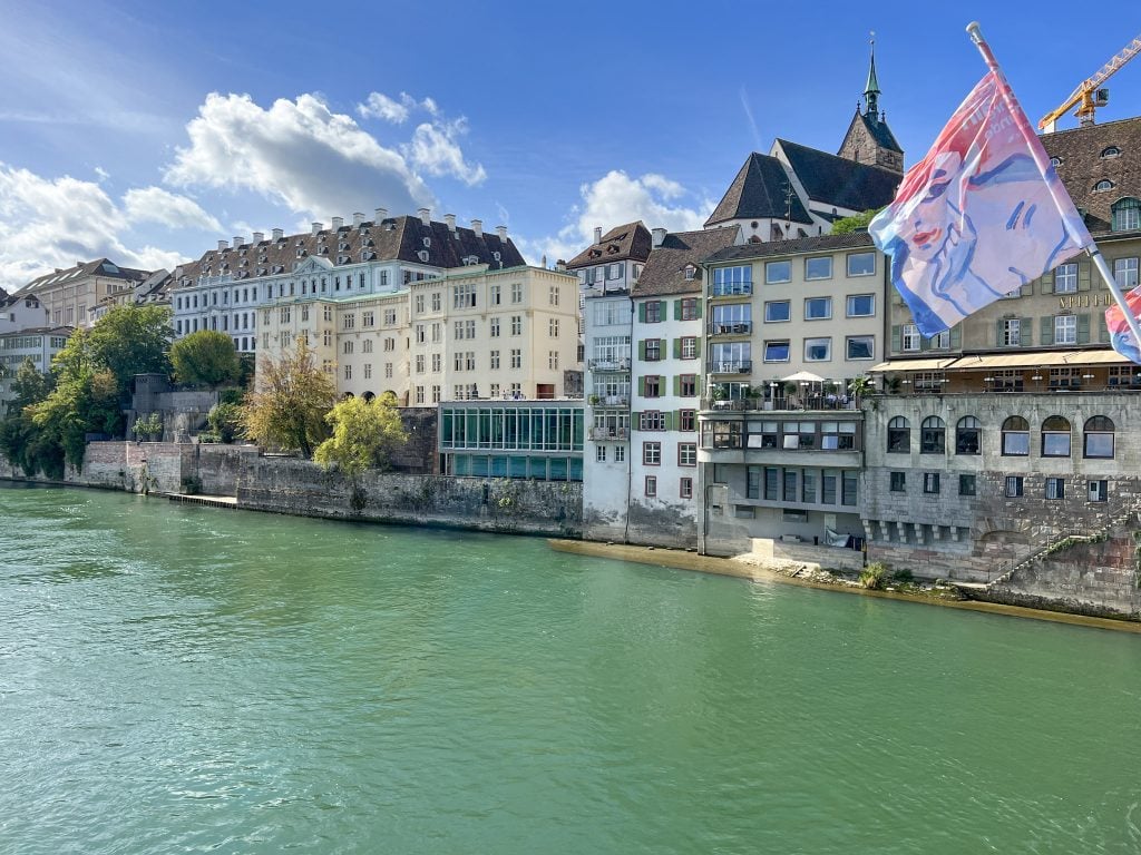 Beautiful view of Basel's riverside with colorful historic buildings and a Swiss flag, showcasing the charming architecture and vibrant atmosphere of the Swiss city.