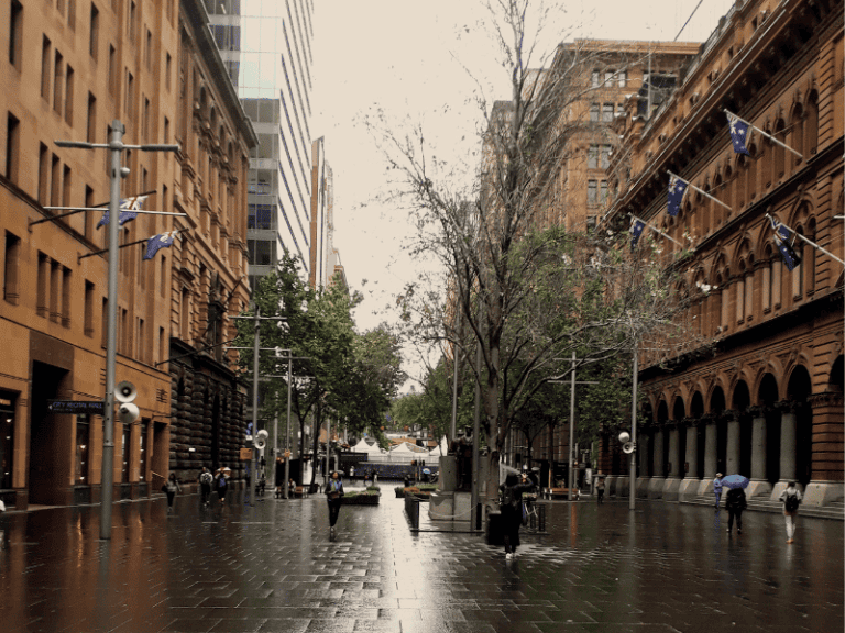 A rainy day in Sydney with people walking under umbrellas along a wet city street, surrounded by historic buildings and trees, capturing the vibrant atmosphere of urban rainy day activities.