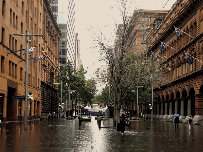 A rainy day in Sydney with people walking under umbrellas along a wet city street, surrounded by historic buildings and trees, capturing the vibrant atmosphere of urban rainy day activities.