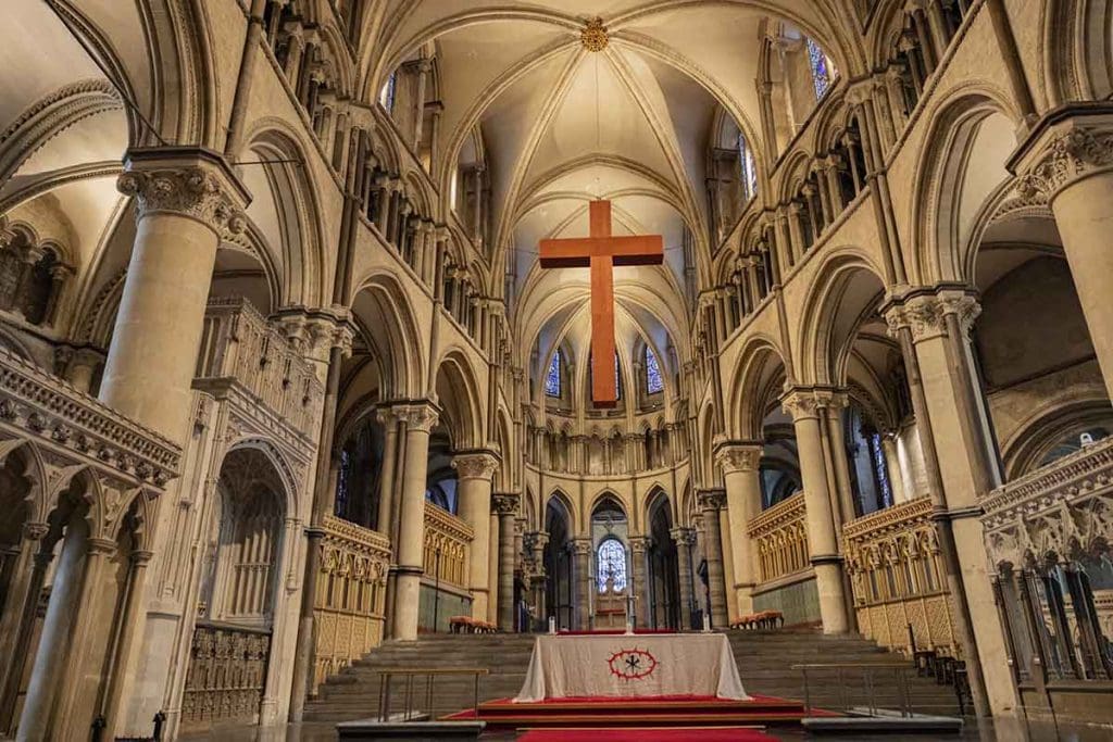 Ancient Gothic church interior featuring soaring arches, intricate stonework, and a prominent wooden cross hanging from the vaulted ceiling, capturing the architectural beauty of East England.