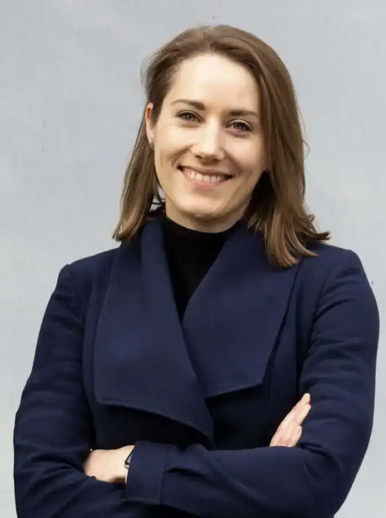 Nina Jameson stands with her arms crossed in front of a grey background and smiles at the camera.