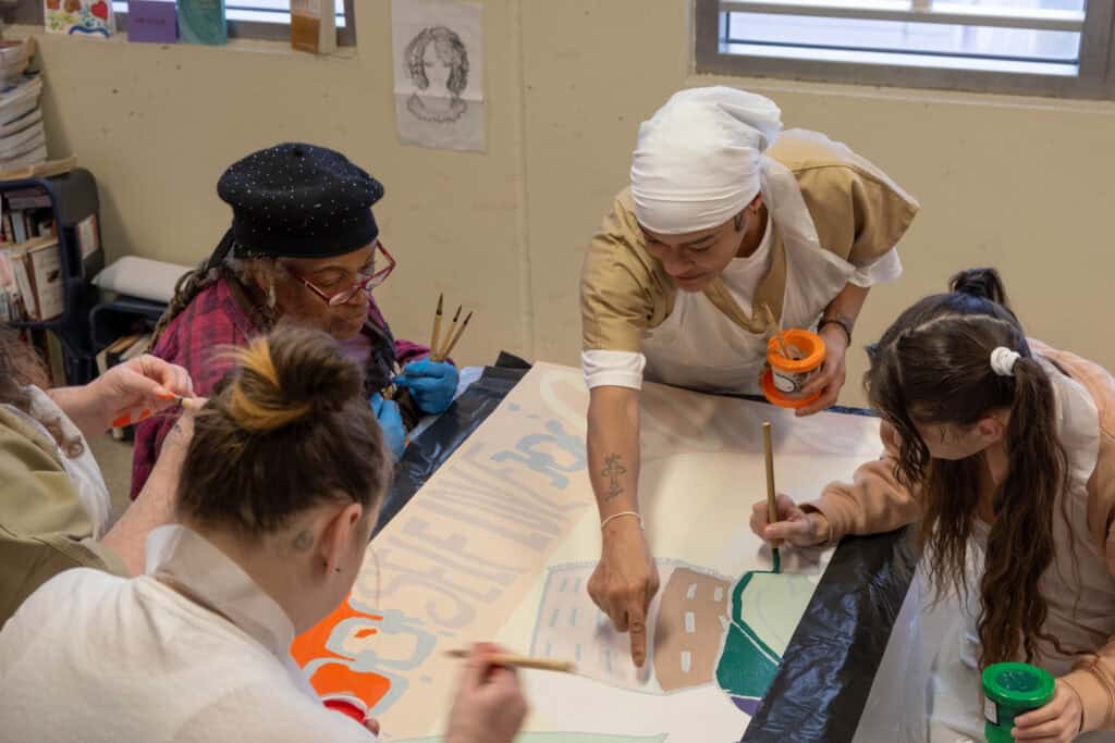 Multimedia artist Dindga McCannon guides NYC Health + Hospitals/Correctional Health Services (CHS) patients as they paint panels of a new mural displayed at the CHS Reentry Service.