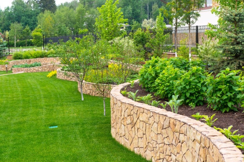 Curved stone retaining wall with lush green lawn and shrubs in a landscaped garden area behind a black fence.