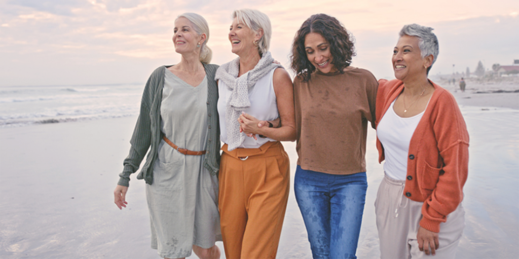 Clinical Trials Norfolk VA | Four women walk along the beach, laughing and smiling.