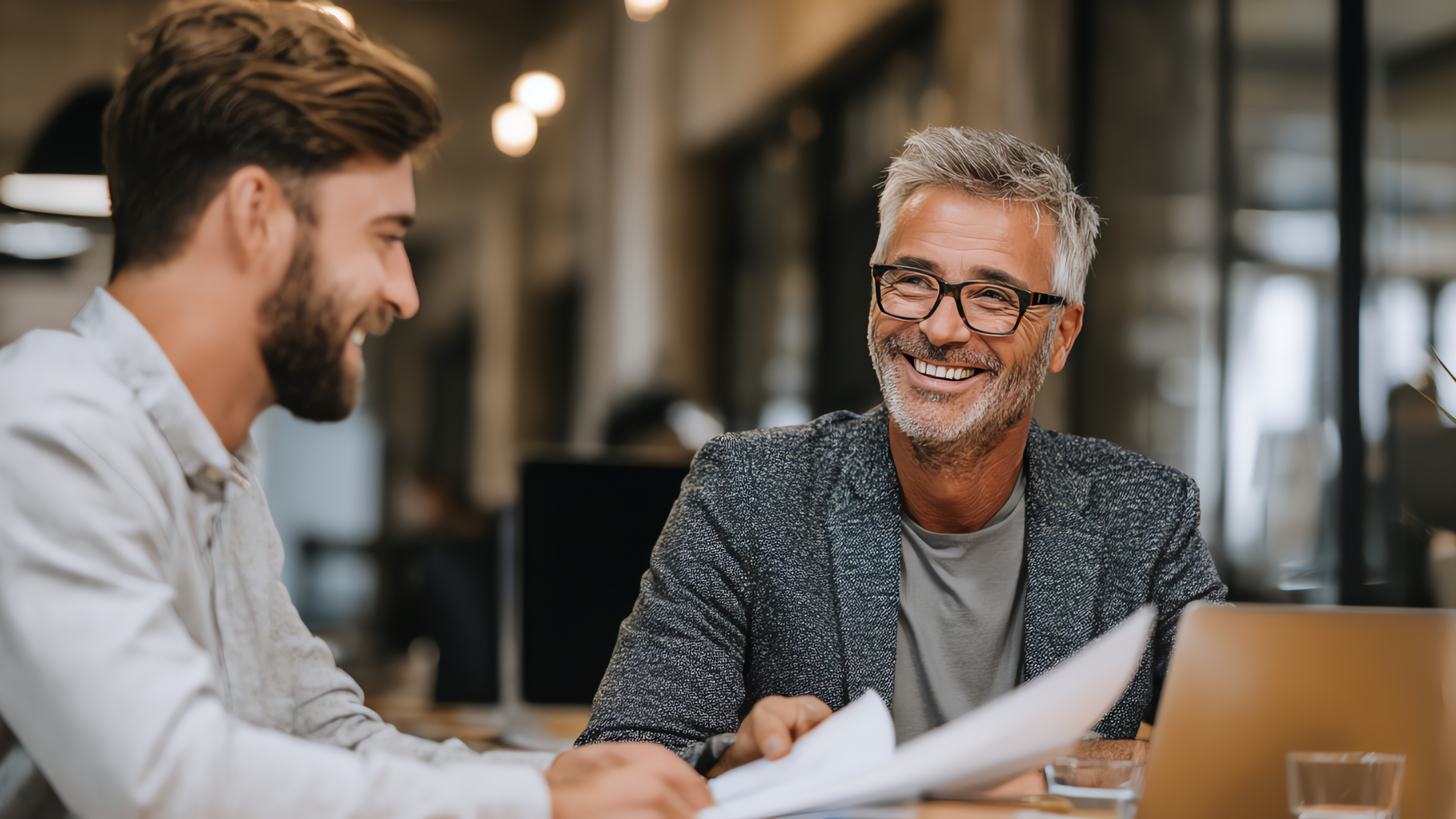 Two businessmen smiling and reviewing loan documents together at a table, discussing SBA loan financing for a business acquisition
