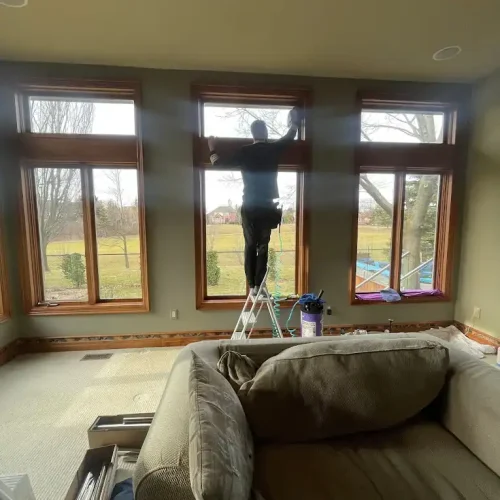A person stands on a stepladder cleaning or working on the upper part of a large window in a living room, with a sofa in the foreground and views of trees and a yard outside.