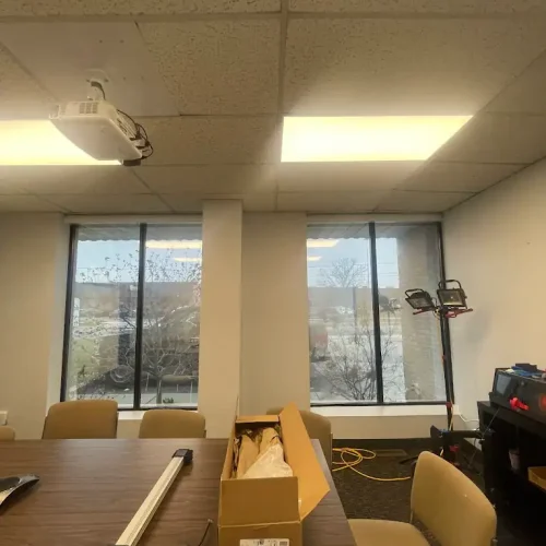 A conference room with beige chairs and a wooden table, on which sits an open cardboard box and a long light fixture. Large windows show an outdoor scene, and a tall work light and cables are in the corner.