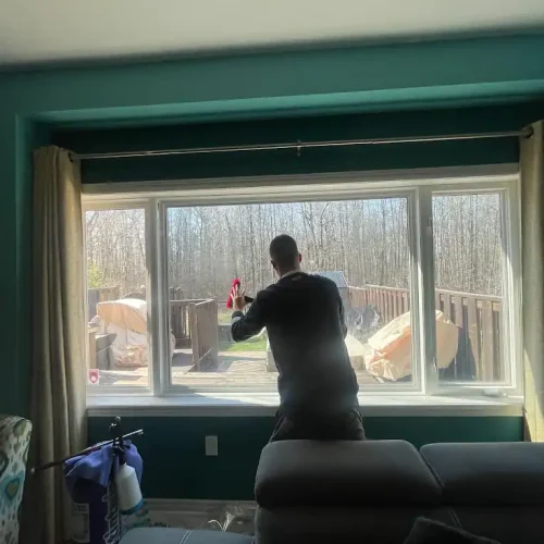 A person stands inside a living room cleaning a large window with a spray bottle. Sunlight streams through the window, revealing a deck and trees outside. Furniture and cleaning supplies are visible indoors.