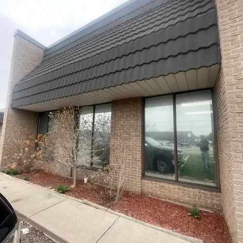 A brick commercial building with three large windows, landscaping with small plants and mulch, and a sidewalk in front. The reflection of a person taking the photo and a parked vehicle are visible in the window.