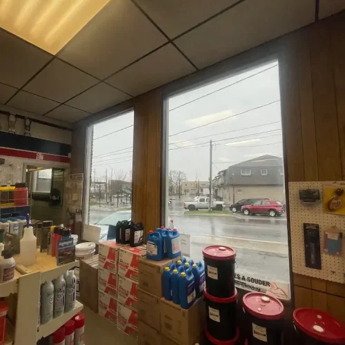A view from inside a store with wooden walls, shelves stocked with cans, boxes, and containers. Large windows show a rainy street, parked cars, and houses outside. Fluorescent ceiling lights are on.