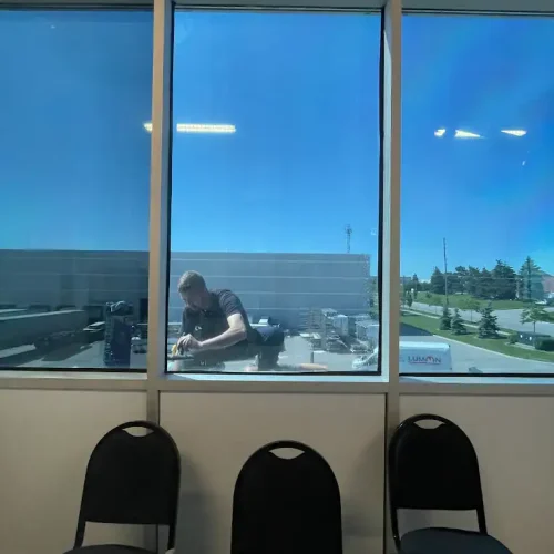 A man is cleaning or working on a ledge outside a large window. Three empty black chairs are positioned inside by the window, and the view includes a clear blue sky and a few trees in the background.