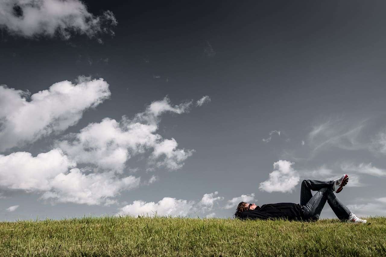 Pensativo joven descansando en un prado bajo cielo nublado en un día soleado.