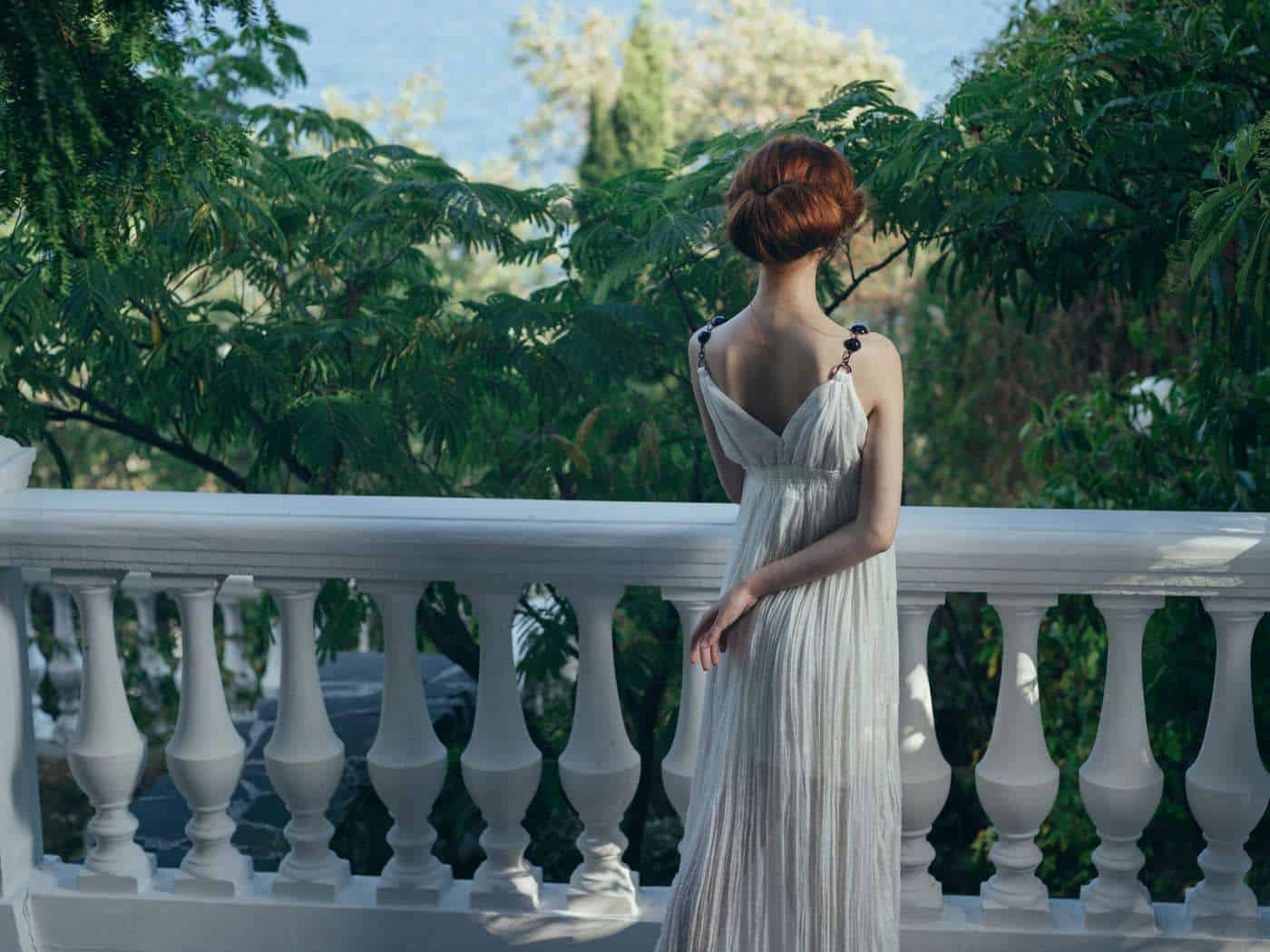 Serena mujer con vestido blanco en balcón con vista a la naturaleza.