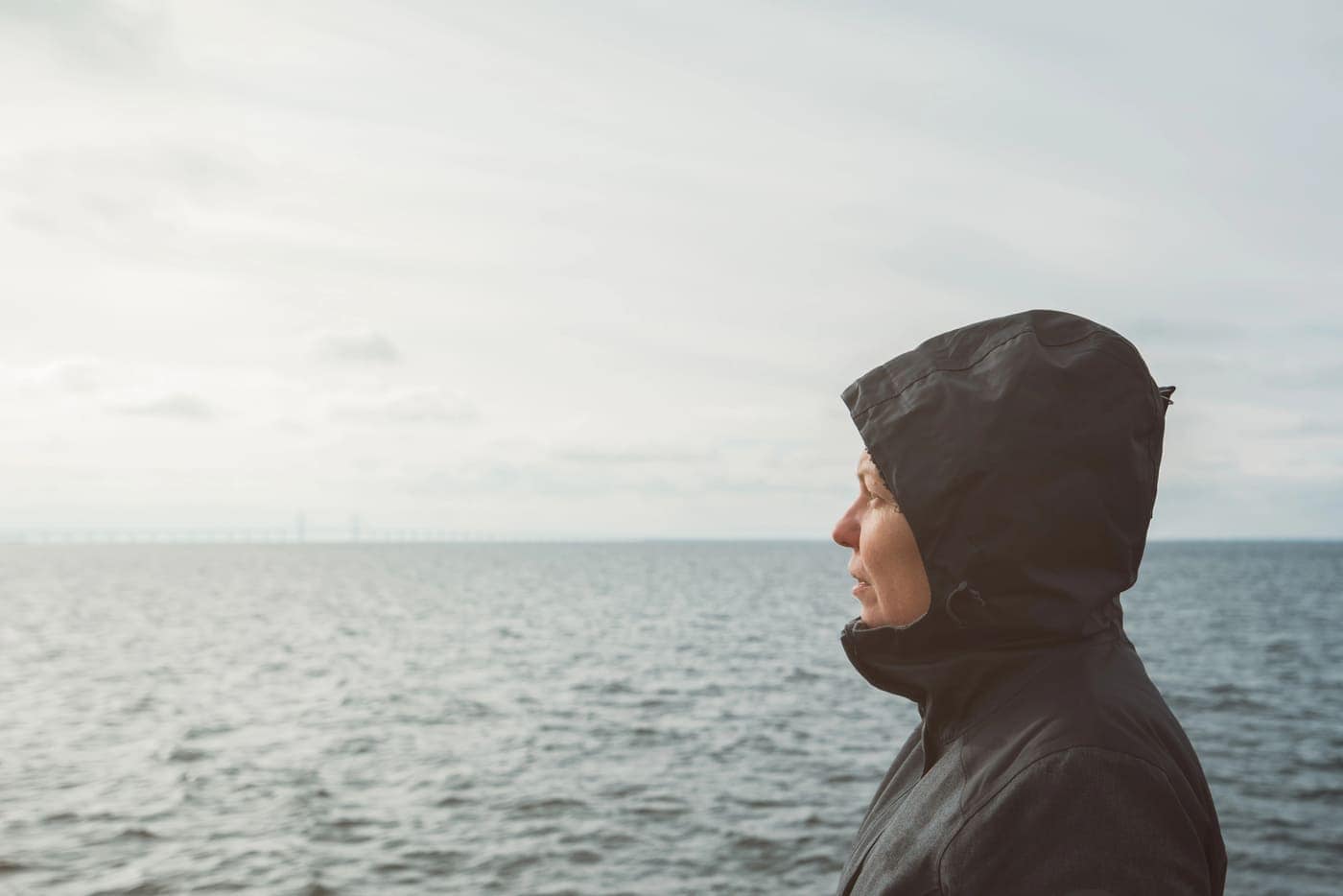 Pensativa mujer con chaqueta y capucha en el mar en un día nublado.